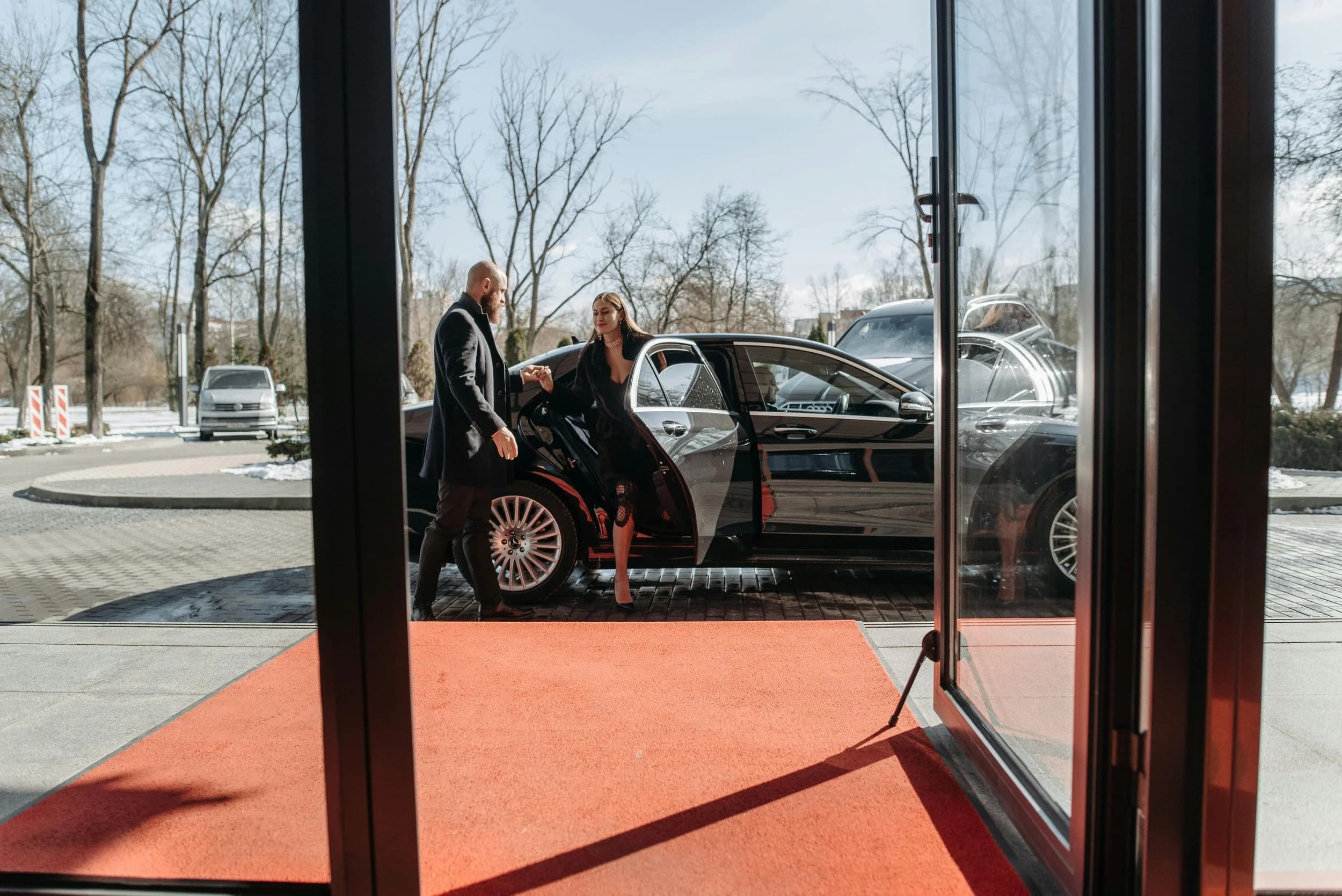 A chauffeur holding the rear door of a black limousine as a guest steps out onto a red carpet